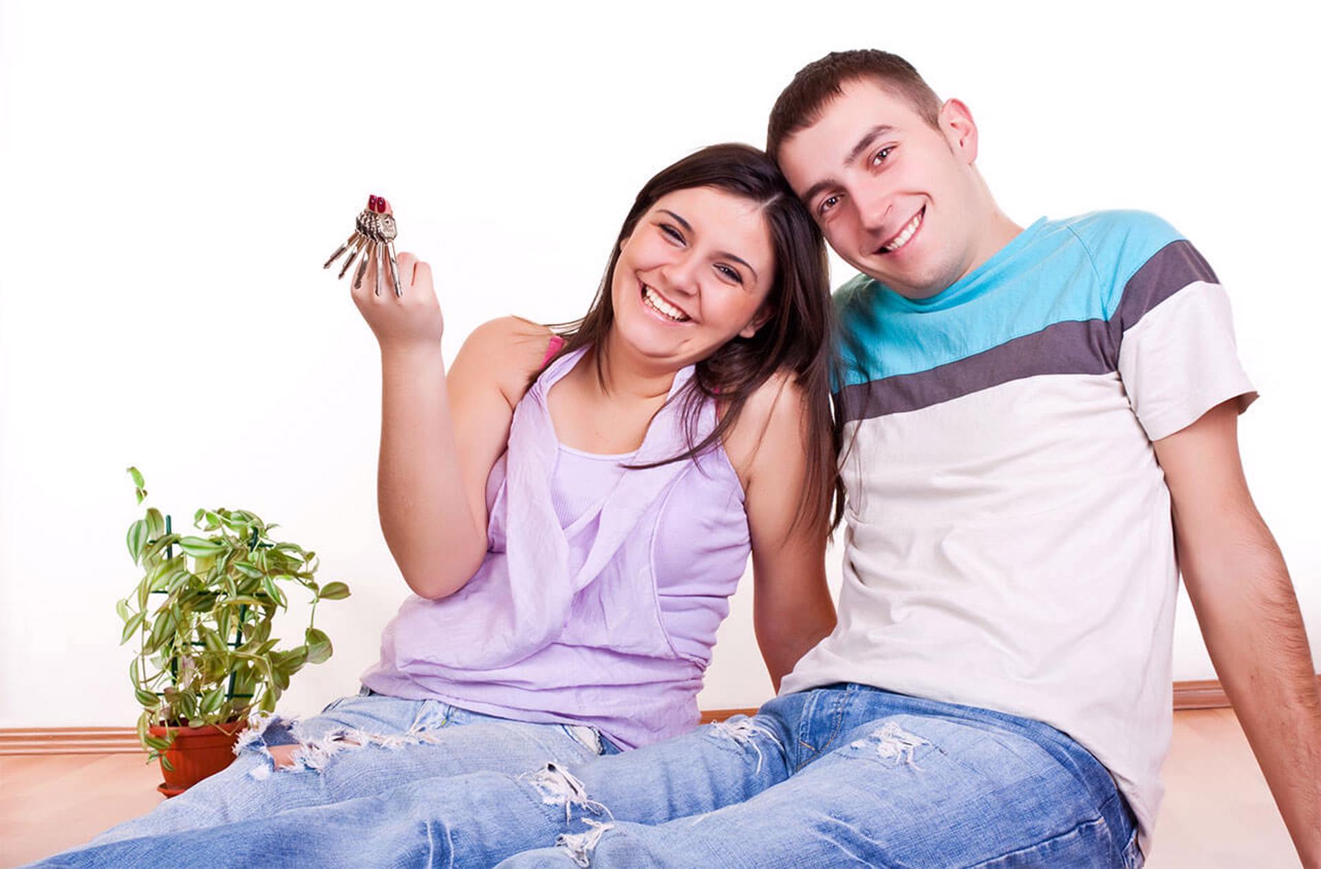 Un jeune couple heureux assis côte à côte avec de grands sourires et tenant des clés de maison.  